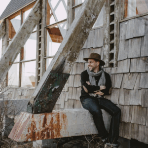 Man in vintage attire sitting by a rustic wooden building.