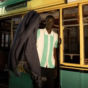 Man standing near a vintage green and yellow tram, wrapped in a dark shawl.
