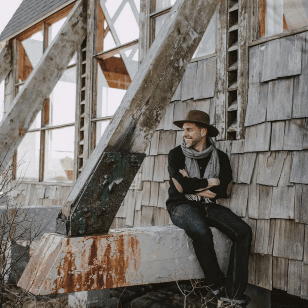 Man in vintage attire sitting by a rustic wooden building.